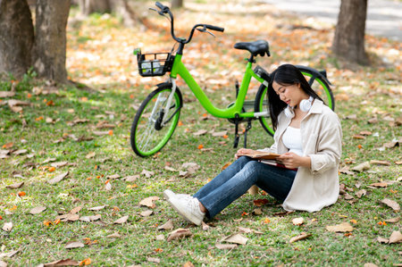 An asian woman is reading the book on her lap while sitting on grass field. Chilling in park, Outdoor activity, Greenery forest park.の写真素材