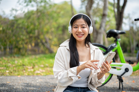 Smiling asian woman is holding smartphone with finger pointing at the screen, with blurred background of bicycle and greenery park. Chilling in the nature.の写真素材