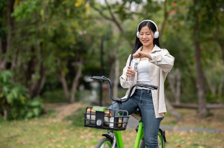 A smiling asian woman is opening a clear water bottle while riding a green bicycle with headphones on. Outdoor activity, Greenery forest park.の写真素材