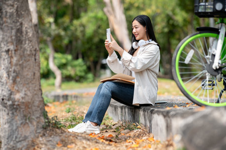 A smiling asian woman is taking a photo with her phone while sitting on the park sidewalk. Taking a selfie or Scenery picture, Chilling outdoor, Greenery forest park.の写真素材