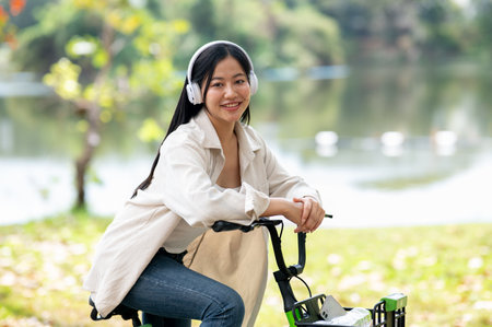An asian woman is putting both hands on the bicycle handlebar while sitting on a bike with headphones on. Bike in park, Outdoor activity, Greenery forest park.の写真素材