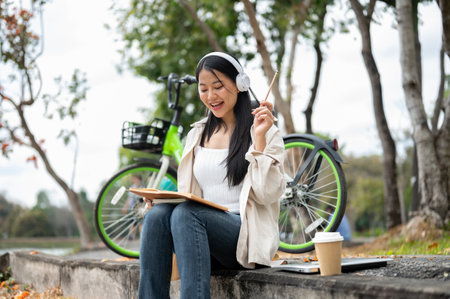 An asian woman is looking happily at her notebook as she figure something out while sitting on the park sidewalk. Study or working outdoor.の写真素材