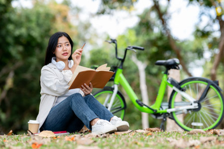 An asian woman is holding a book and pointing away with a pencil while sitting on the park grass field. Chilling in nature, Outdoor activity.の写真素材