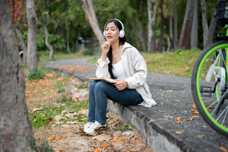 An asian woman is looking ahead while holding her notebook with headphones on, sitting on the park sidewalk. Study or Working outdoor. Greenery forest park.の写真素材
