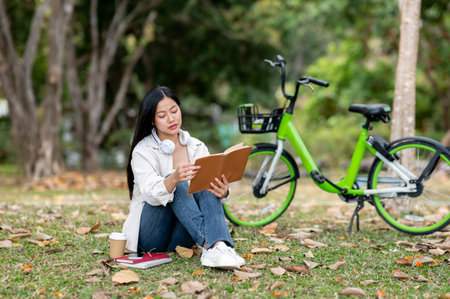 An asian woman is looking intently at the book holding in her hand while sitting on the park grass field. Outdoor activity, Greenery forest park.の写真素材