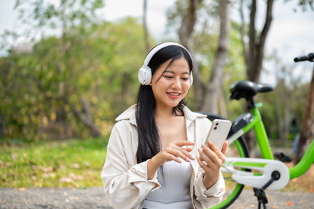 Asian woman is wearing headphones and looking through her smartphone next to the green bicycle. Park cycling. Chilling in the nature.の写真素材