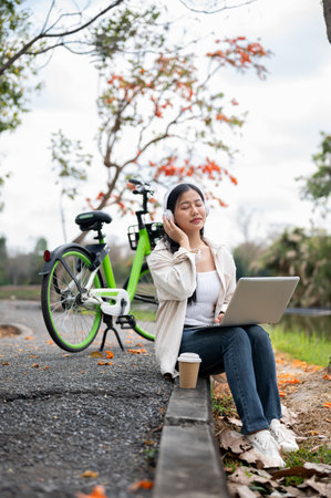 An asian woman is wearing headphones listening to music, sitting on the sidewalk next to her green bicycle. Chilling in the park or garden.の写真素材