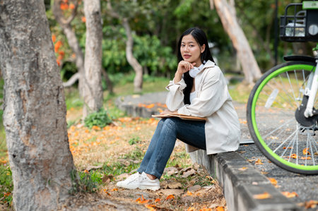 An asian woman is putting a hand under chin and elbow on her notebook while sitting on the park sidewalk. Study or working outdoor. Greenery forest park.の写真素材