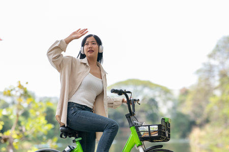 An asian woman is rising a hand to cover the eyes from sunlight while sitting on a bike with headphones on. Bike in park, Outdoor activity, Greenery forest park.の写真素材