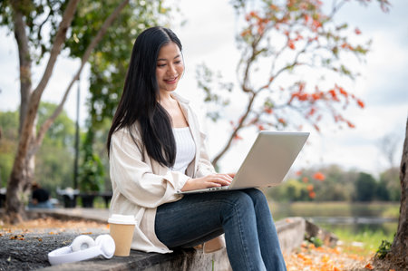An asian woman is looking the laptop on her lap, Working or study outdoor in the park or garden. Blurred background of beautiful nature view.の写真素材