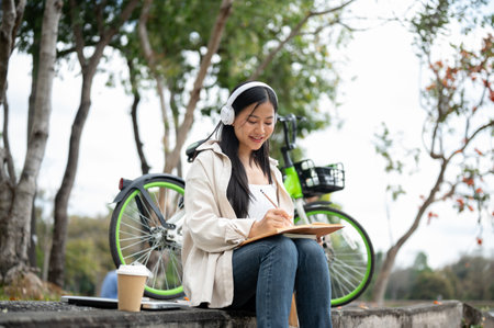An asian woman is happily writing on her notebook with headphones on while sitting on the park sidewalk. Study or working outdoor.の写真素材