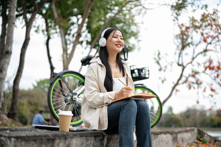 An asian woman is smiling and writing on her notebook with headphones on while sitting on the park sidewalk. Study or working outdoor.の写真素材