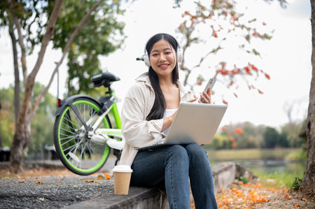 A smiling asian woman is holding smartphone and wearing headphones, sitting alone on the sidewalk next to the green bicycle. Chilling in the park or garden.の写真素材