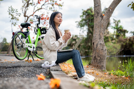 A woman is sitting on the sidewalk and holding a drink with both hand, taking a break from cycling in the park or garden. relaxing in the nature.の写真素材