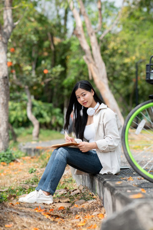 An asian woman is looking straight into camera while writing on her notebook, sitting on the park sidewalk. Study or working outdoor. Greenery forest park.の写真素材