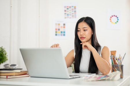 An asian woman is pointing a stylus pencil at laptop screen while putting one hand under chin on table with working materials. Designer working in office.の写真素材