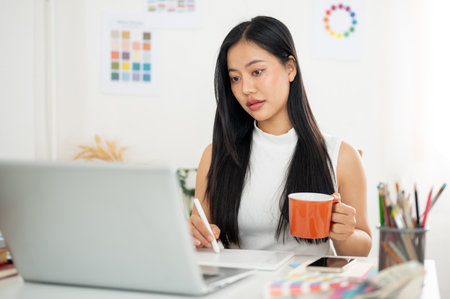 An asian woman is holding a stylus pencil drawing or writing on tablet in one hand and other hand holding a mug. Interior Designer or Graphic Designer working in office.の写真素材