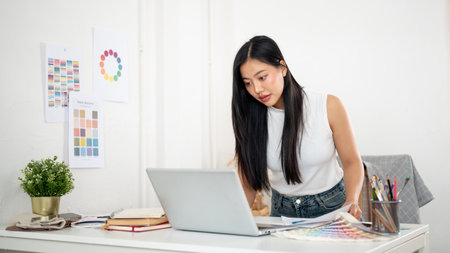 An asian woman is standing and leaning over the table to look closer at the laptop screen. Interior Designer or Graphic Designer in office.の写真素材