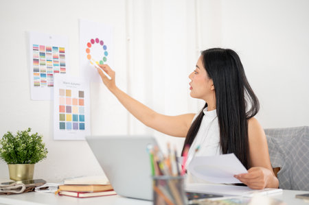 An asian woman is pointing a stylus at a color wheel on wall while holding a document in other hand. Interior Designer or Graphic Designer working in office.の写真素材