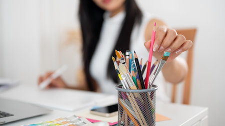 A woman is picking a pink colored pencil from mesh stationary container, sitting at the table with working materials. Interior Designer or Graphic Designer in office.の写真素材
