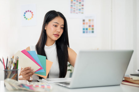 Asian woman is working on the design in laptop with selected color from paper color charts. Interior Designer or Graphic Designer in office.の写真素材