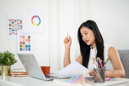 Asian woman is looking at the laptop screen while holding the documents about design color. Interior Designer or Graphic Designer working in office.の写真素材