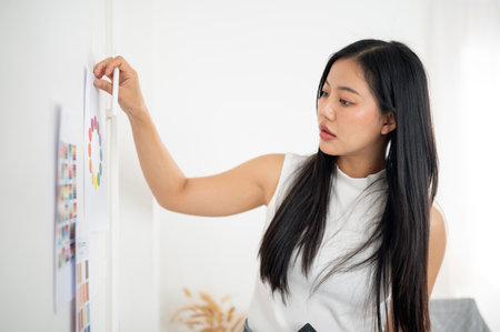 Asian woman is sticking a printed color charts or a color wheel on wall while holding a stylus. Interior Designer or Graphic Designer working in office.の写真素材