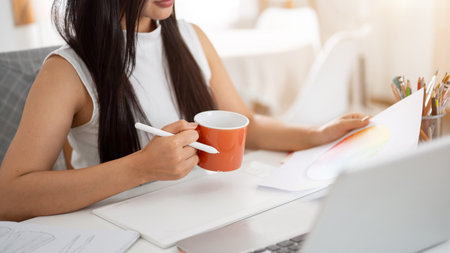 A close up of a hand holding an orange ceramic mug with a stylus while reading through the document. Interior Designer or Graphic Designer in office.の写真素材