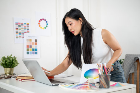 An asian woman leaning over the table to look at laptop screen while holding a stylus and printed color chart. Interior Designer or Graphic Designer in office.の写真素材