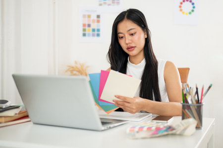 An asian woman is picking the color from paper color charts to use in her design, sitting with laptop at working table. Interior Designer or Graphic Designer in office.の写真素材