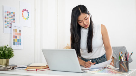 An asian woman standing and leaning over the table to read the documents placing among other working materials. Interior Designer or Graphic Designer in office.の写真素材