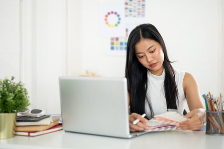An asian woman is looking and choosing color from the printed color charts or color palettes in her hand. Interior Designer or Graphic Designer working in office.の写真素材