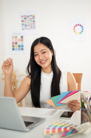 Asian woman is smiling while holding the selected color from paper color charts. Interior Designer or Graphic Designer working in office.の写真素材