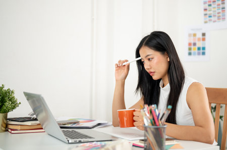 An asian woman is thinking thoughtfully while looking at laptop with a mug and a stylus in each hand. Designer in office.の写真素材