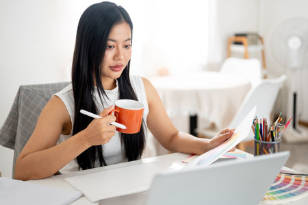 An asian woman is holding an orange ceramic mug with a stylus while looking at the laptop screen. Interior Designer or Graphic Designer in office.の写真素材
