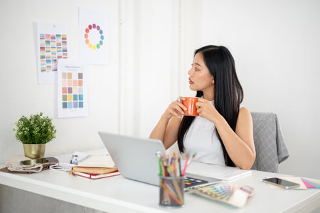 Asian woman is looking at a color wheel or color palette on wall while holding a mug with both hand. Interior Designer or Graphic Designer working in office.の写真素材