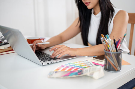 An asian woman is holding a stylus and typing on laptop, sitting at the table with working materials. Interior Designer or Graphic Designer working in office.の写真素材