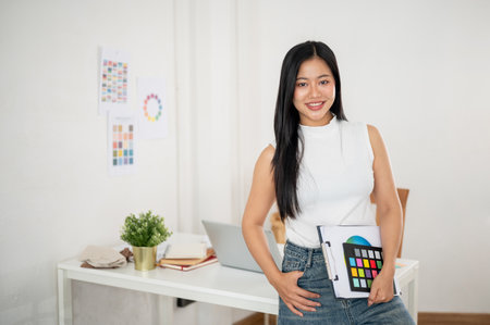 A smiling asian woman is carrying the clipboard in one hand while posing in front of her work desk. Interior Designer or Graphic Designer in office.の写真素材