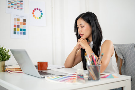 An asian woman is putting both hand under her chin and staring intently at laptop screen on the table. Interior Designer or Graphic Designer working in office.の写真素材
