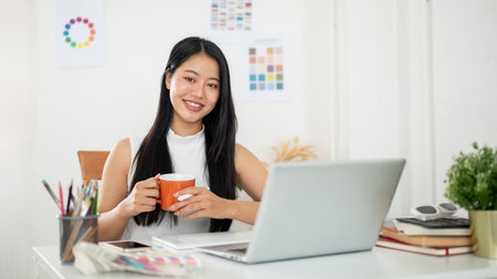 A smiling asian woman is holding a mug with both hands, sitting in front of a laptop with working materials on table. Interior Designer or Graphic Designer in office.の写真素材