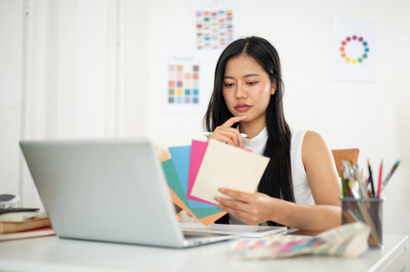 Asian woman is thinking deeply while choosing the color from paper color charts, sitting with a laptop at the working table. Interior Designer or Graphic Designer in office.の写真素材