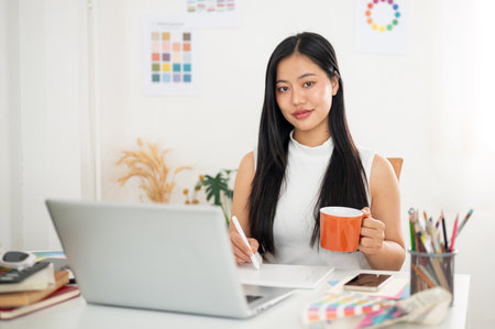An asian woman is holding a stylus pencil drawing or writing on tablet in one hand and other hand holding a mug. Interior Designer or Graphic Designer working in office.の写真素材