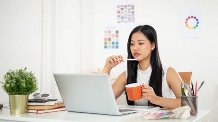 An asian woman is thinking thoughtfully while holding a mug, sitting in front of a laptop with working materials on table. Interior Designer or Graphic Designer in office.の写真素材