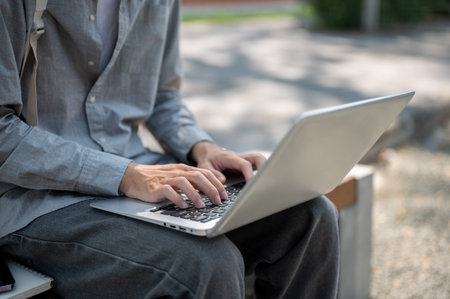 Close up of a pair of hands in grey shirt typing or chatting on the laptop, sitting on sidewalk bench. Spending free time, Study or working outdoor.の写真素材