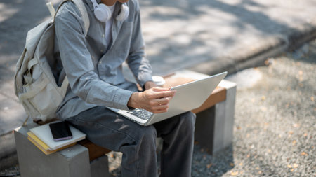 Close up of an asian man touching or adjusting his laptop screen, sitting on the sidewalk bench. Spending free time, Study or working outdoor.の写真素材
