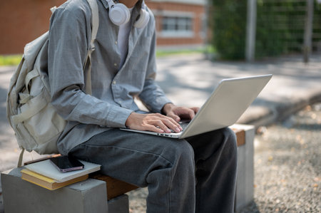 Close up of a man in grey shirt typing or chatting on his laptop, sitting next to the books and a phone on sidewalk bench. Spending free time, Study or working outdoor.の写真素材