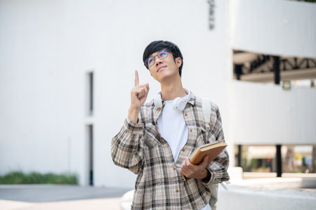 An asian man with glasses is holding books and pointing a finger up while standing in front of a building. Thinking thoughtfully or figuring thing out.の写真素材