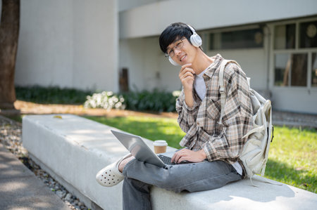 A smiling asian man with glasses is listening to music in his headphones while sitting with laptop on concrete bench in front of building. Chilling Outdoor.の写真素材