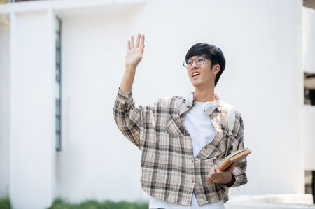 An asian man with glasses is holding books and waving a hand while standing in front of a building. Greeting or send off someone.の写真素材