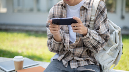Close up of a man looking at smartphone while sitting with coffee and books on concrete bench in park. Study or Working Outdoor.の写真素材